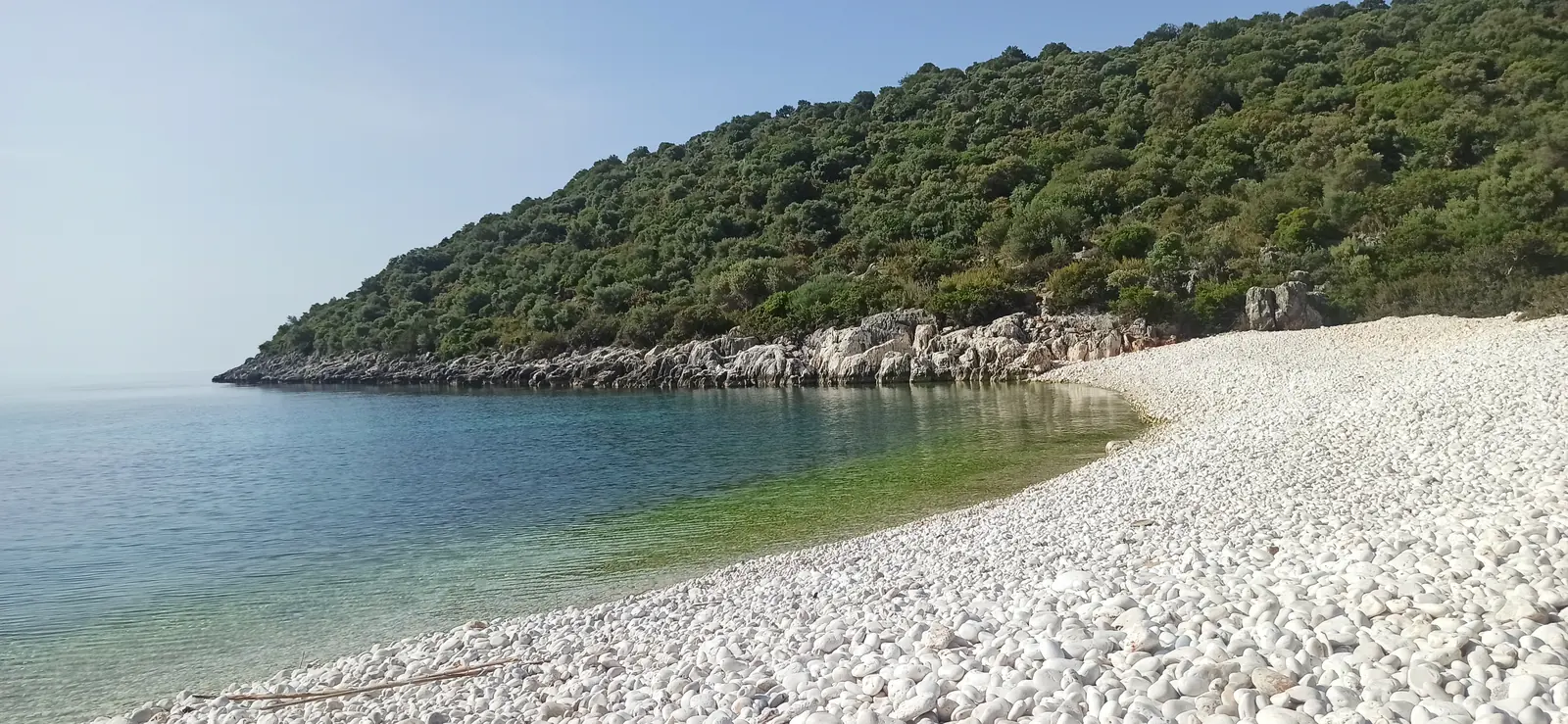 A white pebble beach curving around clear green water below a wooded hill