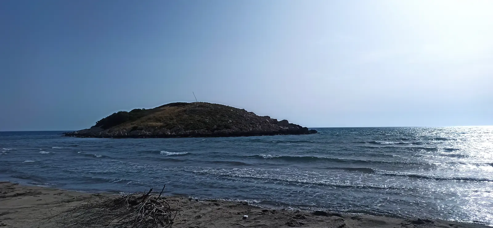 A low dark island offshore, waves rolling onto a beach under a hazy sun