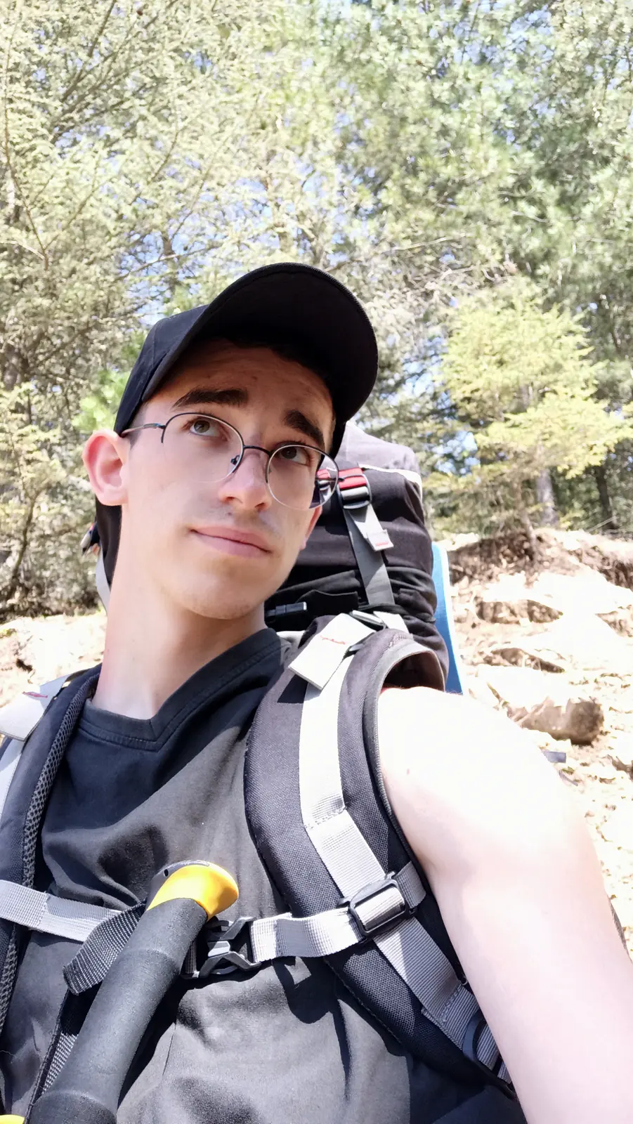 Andrii in a cap and glasses, wearing a large backpack, among pines and rocks
