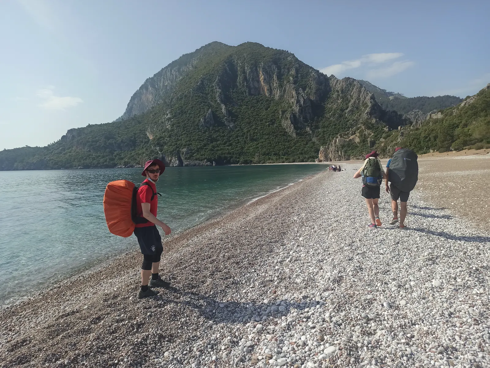 Hikers on a pebble beach, one with an orange backpack, a steep mountain behind