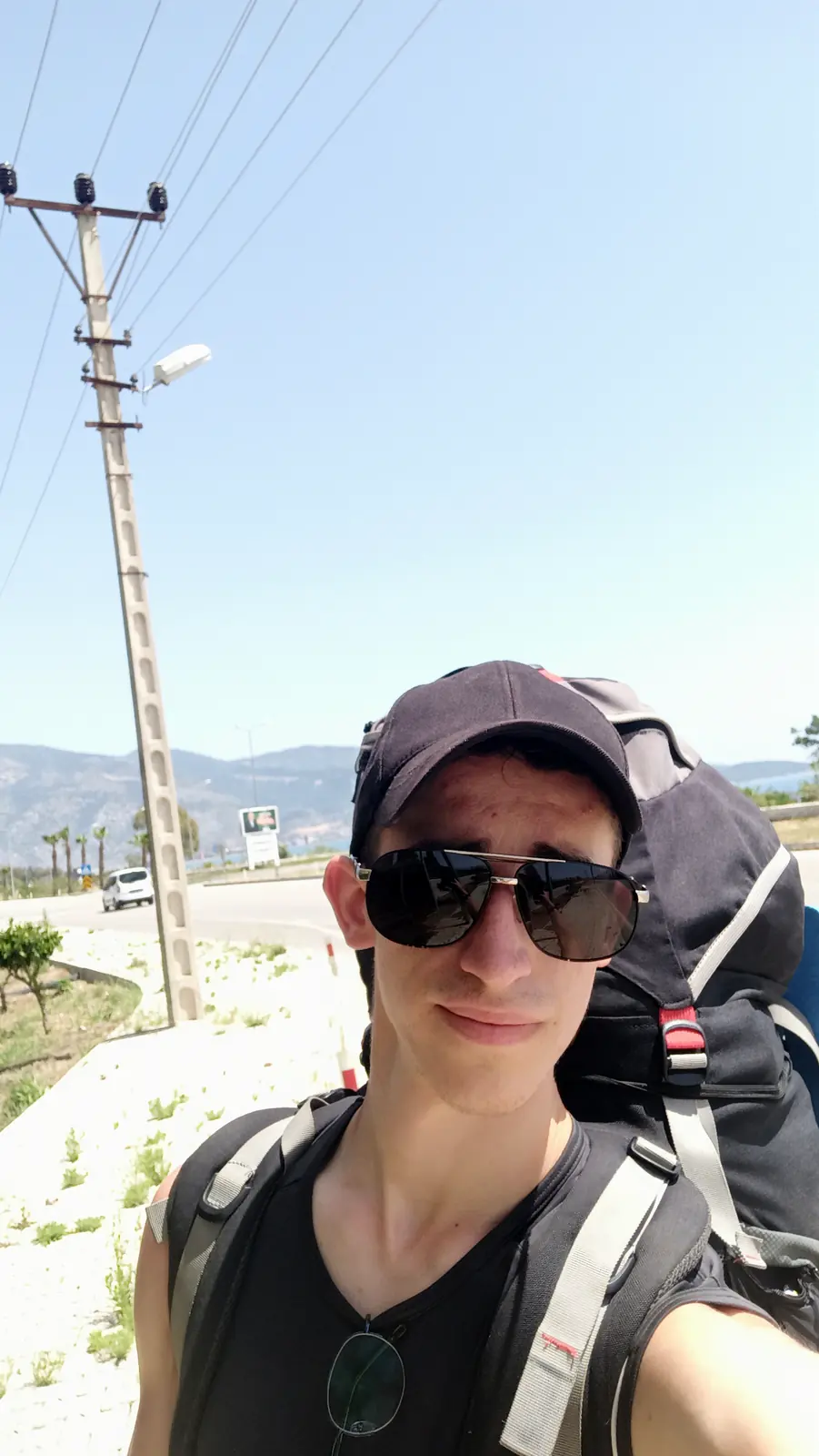 Andrii in sunglasses and a cap with his backpack, beside a road under power lines