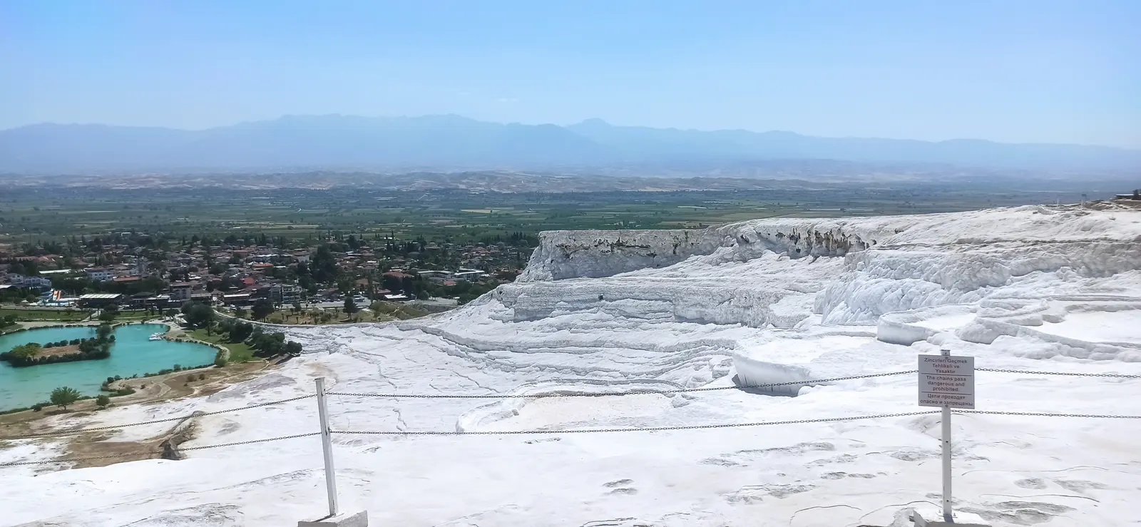 White terraces above a town with a turquoise pond and a wide plain beyond